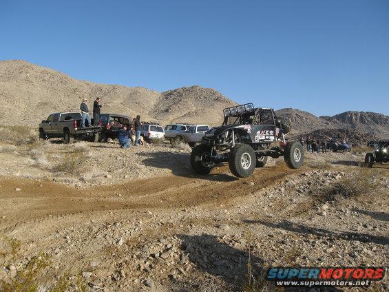 Buggy kicking up dirt on a desert trail with people and other vehicles watching