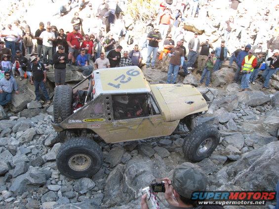 Off-road buggy with number 927 on top, navigating a rocky trail with a crowd watching