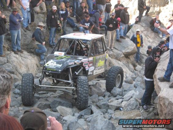Off-road buggy with large tires navigating a rocky terrain, surrounded by spectators