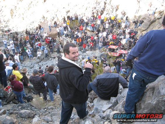 Red off-road vehicle tilted on rocky terrain with a crowd of spectators watching
