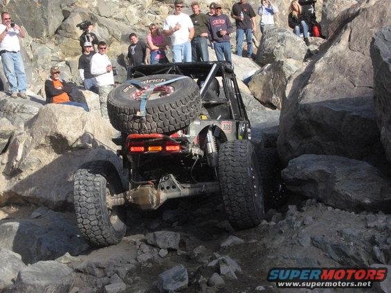 Off-road vehicle with spare tire mounted on rear, climbing over large rocks. Spectators watch from above
