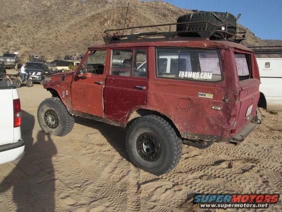 Red off-road SUV with a roof rack and spare tire on top, parked in sand with other vehicles around