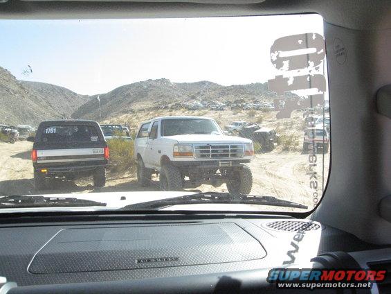 White Ford Bronco with lifted suspension and off-road tires, parked on a dirt road with other vehicles in the background
