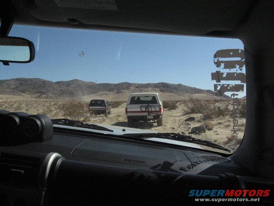 White SUV with a bike rack on the back and a dark SUV behind it driving on a dirt road in a desert