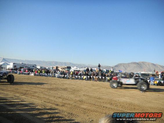 Off-road buggy races past spectators lined up behind a fence. Another buggy is visible on the left