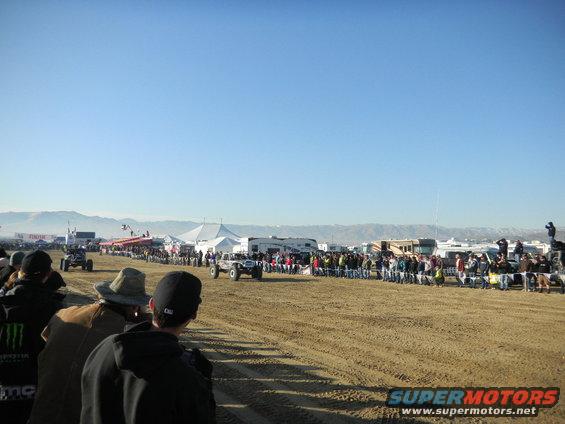 Off-road vehicles race past spectators in a sandy area under a clear blue sky