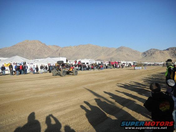 Off-road buggy with roll cage and spare tire drives on dirt track, spectators and tents in background