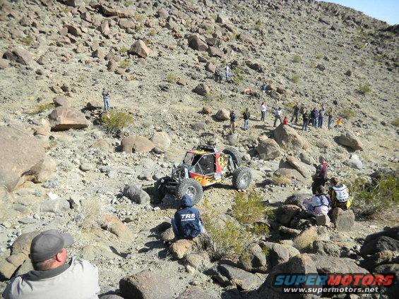 Off-road buggy with flame decals climbing a rocky hill with spectators watching