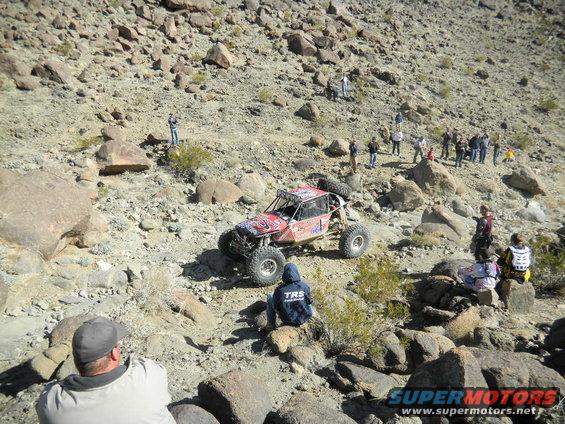 Red off-road buggy navigates rocky terrain with spectators watching