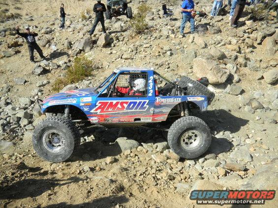 Off-road race truck with AMSOIL livery climbing a rocky hill; another Jeep is visible in the background