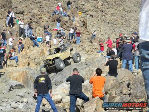 Off-road vehicle with large tires climbing a rocky slope, watched by a crowd