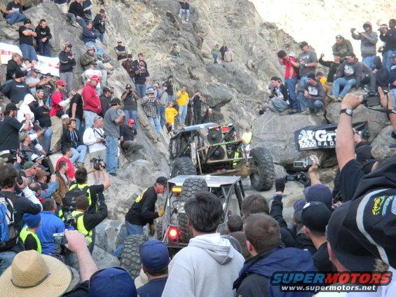 Off-road buggy navigates a steep, rocky incline as a crowd watches from the hillside