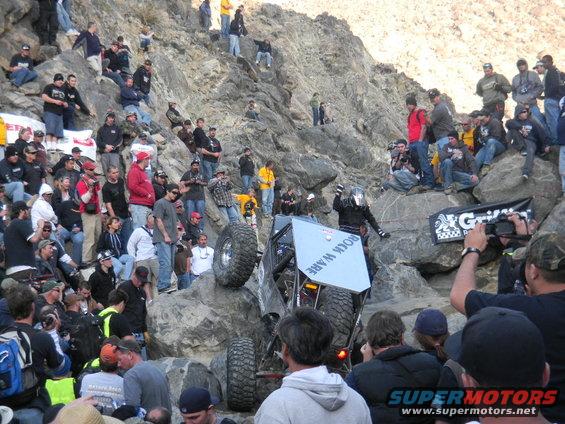 Off-road vehicle with large tires climbing a rocky incline, surrounded by spectators