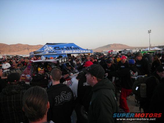 Crowd gathered around off-road race vehicles under a blue tent with '1-800-RACERAD' on it. Desert mountains in background