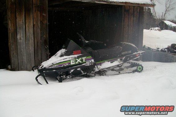 Black 1990 Arctic Cat El Tigre EXT snowmobile covered in snow, parked next to a wooden structure