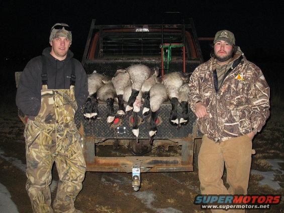 Two guys stand behind a truck bed full of dead geese. One guy wears camo overalls, the other a camo jacket