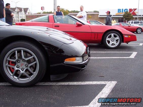 Black Corvette in foreground, red Corvette behind it in parking lot. People standing around