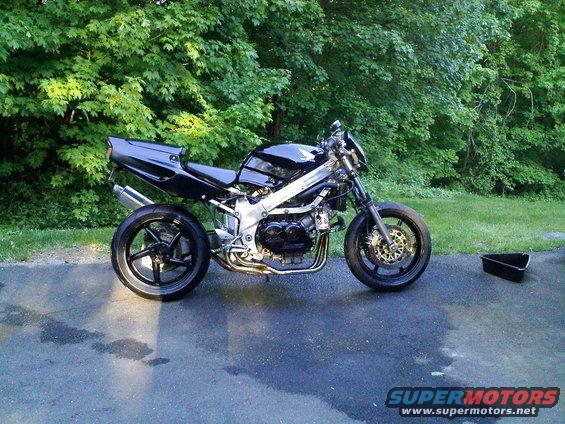Black motorcycle with exposed engine and exhaust pipes, parked on asphalt in front of trees