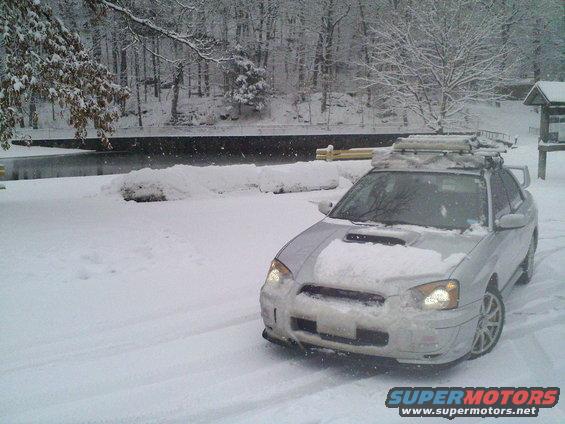 Silver Subaru Impreza WRX with a roof rack covered in snow, parked in a snowy area near a body of water