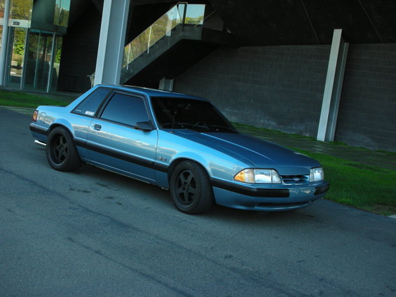 Blue 1990 Ford Mustang coupe parked on asphalt with a modern building in the background