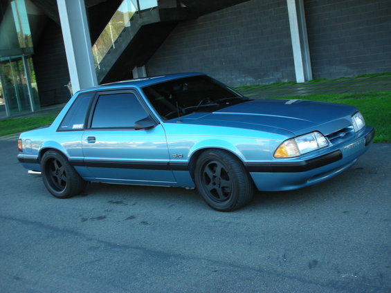 Blue 1990 Ford Mustang coupe parked on asphalt, with a modern building and stairs in the background