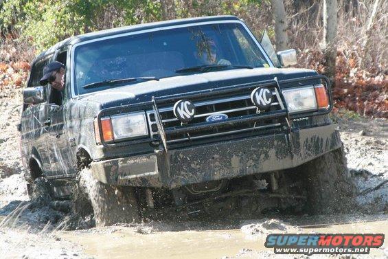 Black Ford Bronco driving through mud, KC lights on grille guard