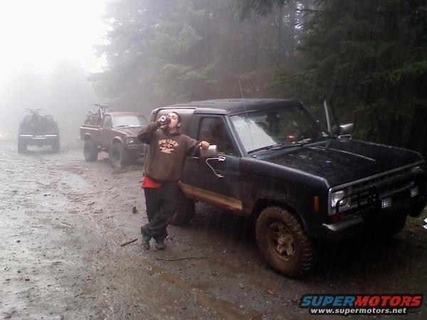 Black 1984 Ford Bronco II parked on a muddy dirt road with a person drinking from a bottle leaning on it