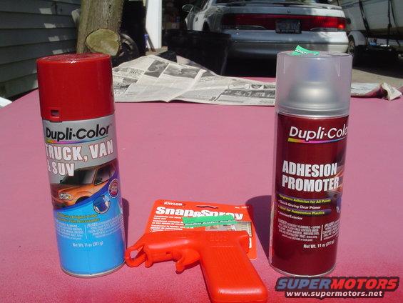 Two Dupli-Color spray cans and a red spray trigger on a pink surface, with a silver sedan in the background