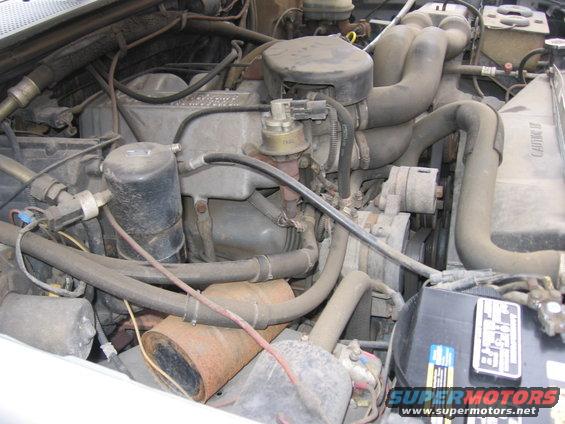 Engine bay with hoses, belts, and engine components visible. A battery is in the lower right