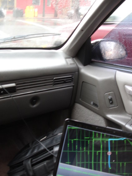 1989 Ford Taurus interior, gray dashboard, laptop displaying map, black briefcase, rain on windshield, red car visible outside