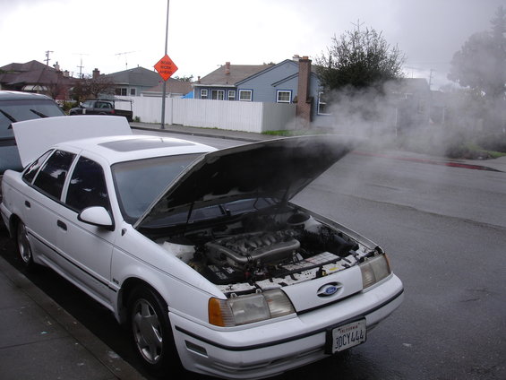 White sedan parked on street, hood up, engine exposed, steam billowing out. License plate reads 3DCY444