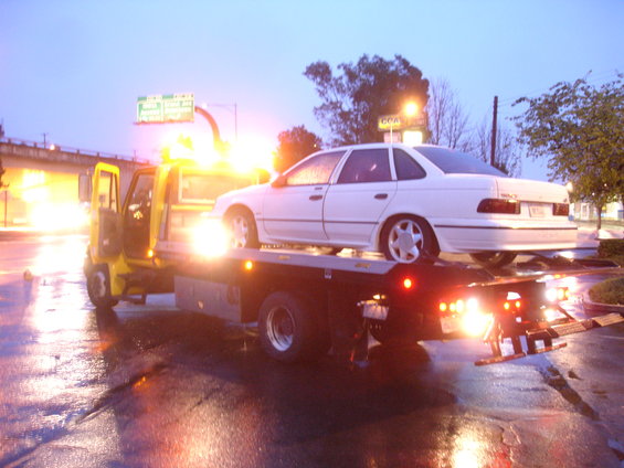 White 1989 Ford Taurus sedan being towed on a flatbed tow truck at dusk