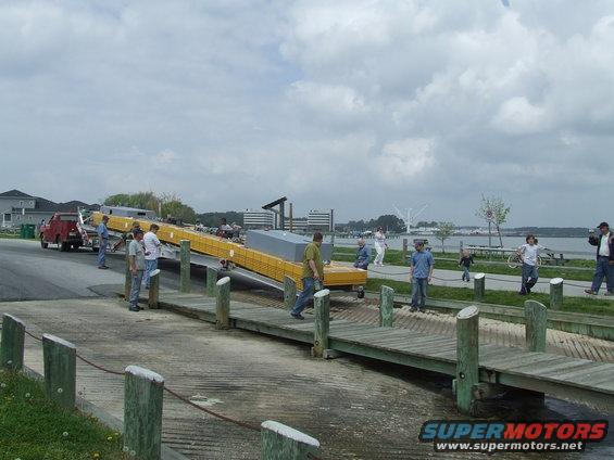 People gather around a boat ramp and dock, with a trailer and a red truck visible in the background
