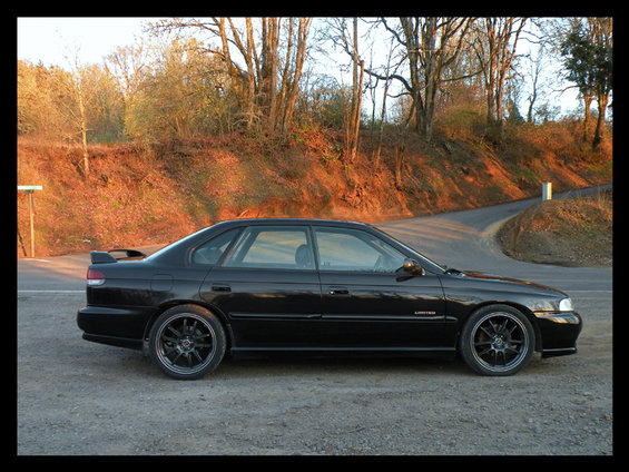 Black 1998 Subaru Legacy sedan with a rear spoiler and aftermarket wheels, parked on a gravel surface