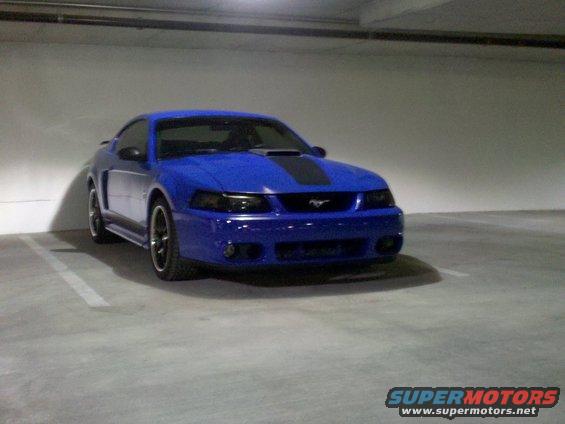 Blue Ford Mustang with black racing stripes parked in a garage, front 3/4 view