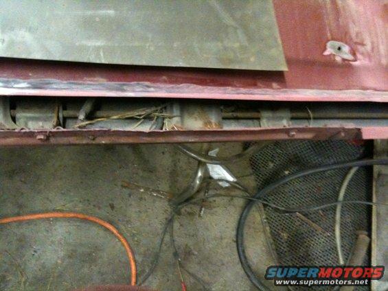 Undercarriage of a red vehicle showing rusted metal, wires, and a black mesh grate