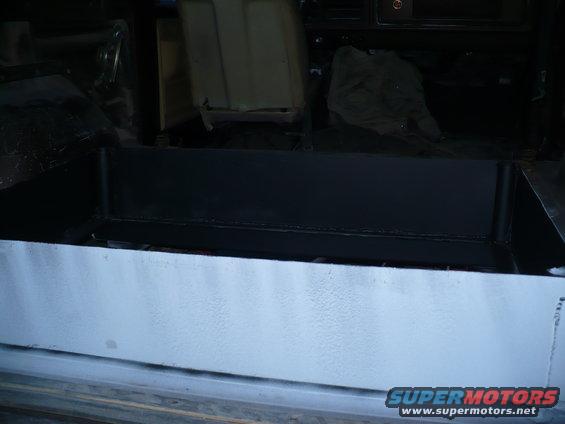 Vehicle interior cargo area with black metal box and light gray painted sides. Some seats are visible in the background