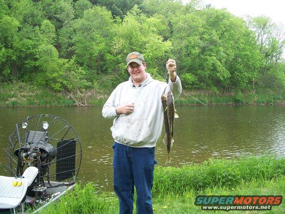 Guy holding two fish next to an airboat on a riverbank
