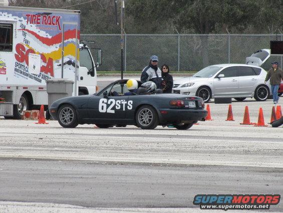 Dark convertible sports car with '62STS' on the side, parked near a trailer and a silver hatchback