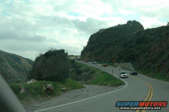 Several cars driving on a two-lane road that winds through a hilly, green landscape under a cloudy sky