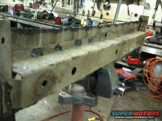 Rusted metal frame part held in vise, showing weld marks and holes. Tools and equipment visible in background