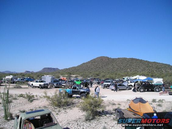 Desert scene with many trucks and cars parked, some with tents, under a clear blue sky
