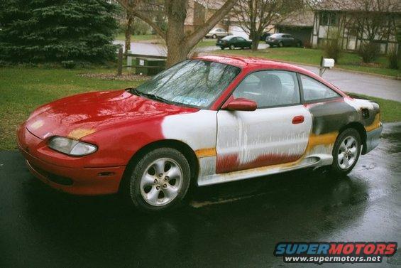 Red and white 1994 Mazda MX-6 coupe with primer patches and rust, parked on wet asphalt