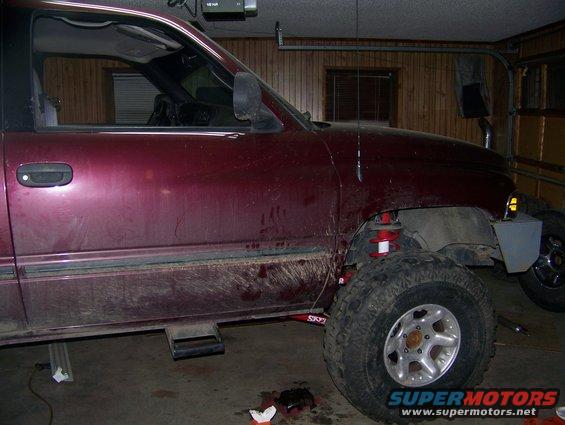 Maroon Dodge pickup truck, side view, lifted with red shocks. It's in a garage