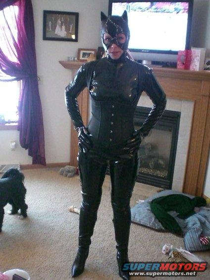Person in a shiny black catwoman costume with mask and ears, standing in a living room with a dog and a fireplace