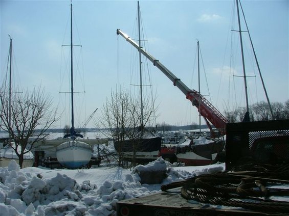 Boats covered in snow at a marina, with a large crane positioned over them. Bare trees and a bright sky are visible