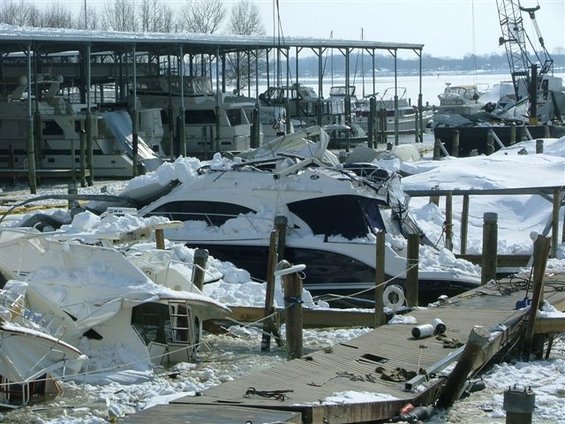 Boats in a marina are damaged and covered in snow. Some boats are overturned or partially submerged