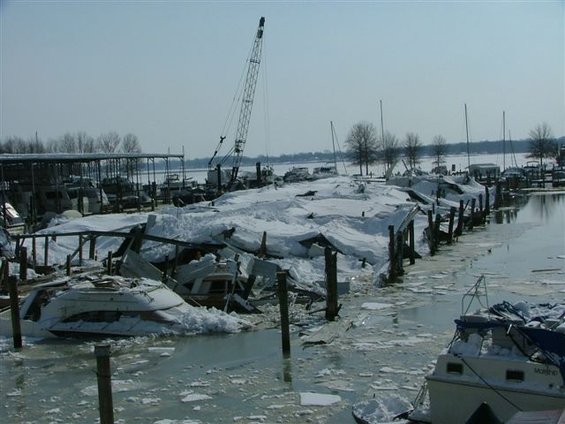 Boats and docks damaged by snow and ice at a marina. A crane is visible in the background