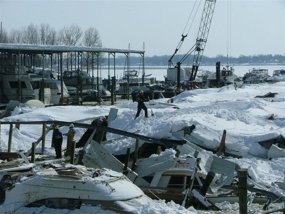 Boats damaged and covered in snow at a marina, with a crane and people working