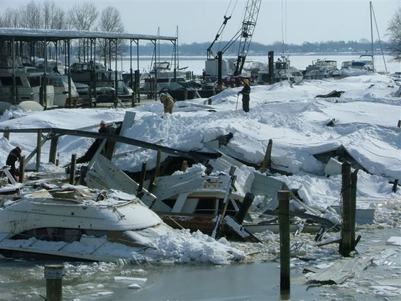 Damaged boats covered in snow and ice at a marina, with a crane in the background